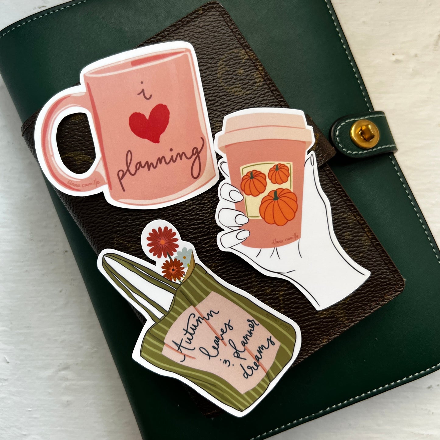 Three die cut planner stickers, one featuring a pink mug with the writing I love planning, a sticker with a hand holding a pumpkin spice latte coffee cup, a sticker of a tote bag that is green with green stripes and flowers sticking out of it with the words "Autumn leaves & planner dreams" 